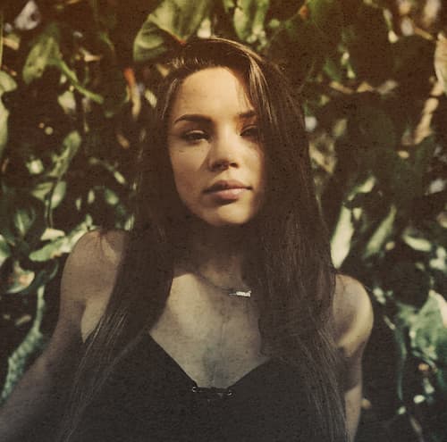 Brunette standing under trees with shadows all over looking into camera